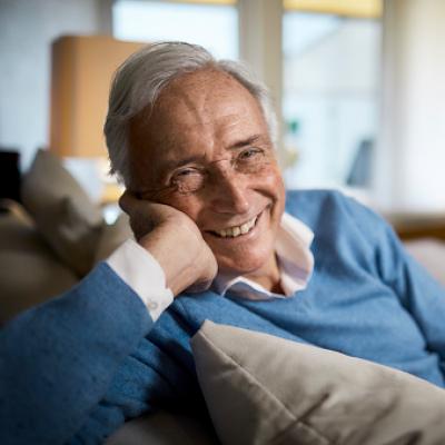 A gray haired gentleman smiling, looking at you by leaning over the back of his couch